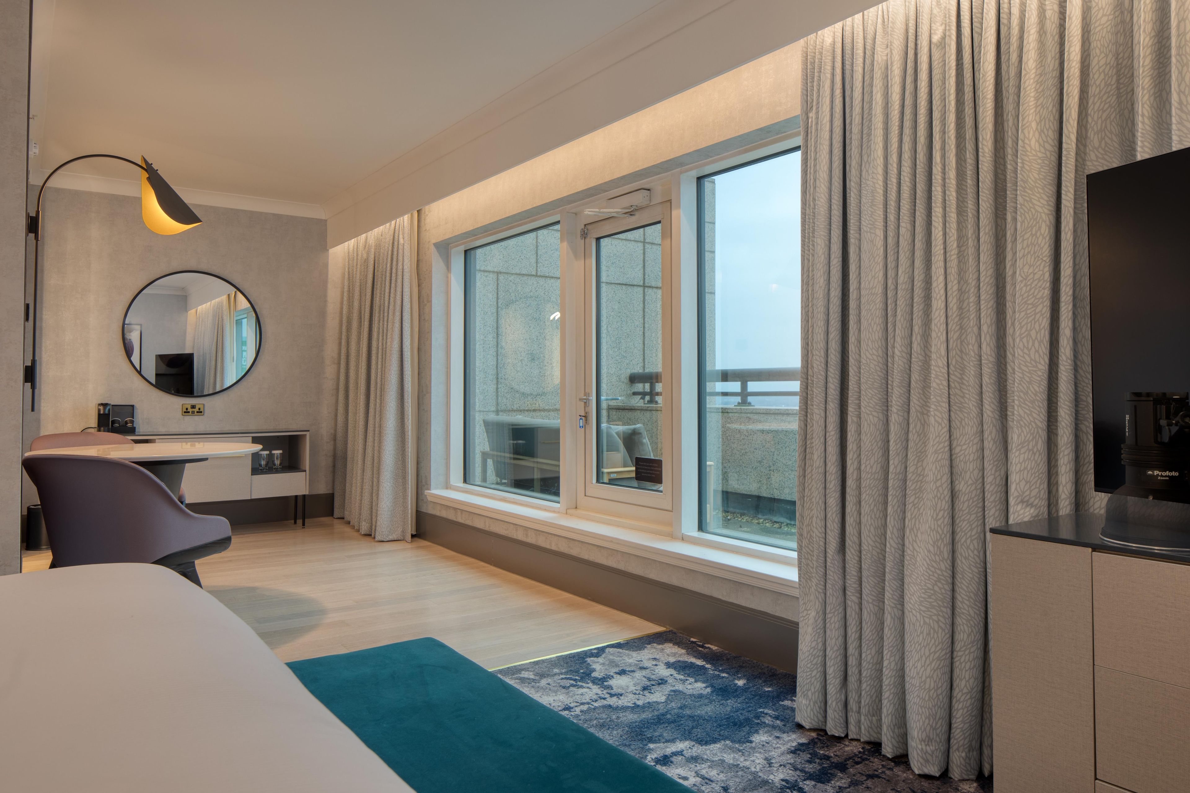 A hotel room with a bed, a desk, a chair, a round mirror, a window with curtains, and a view of the outside.