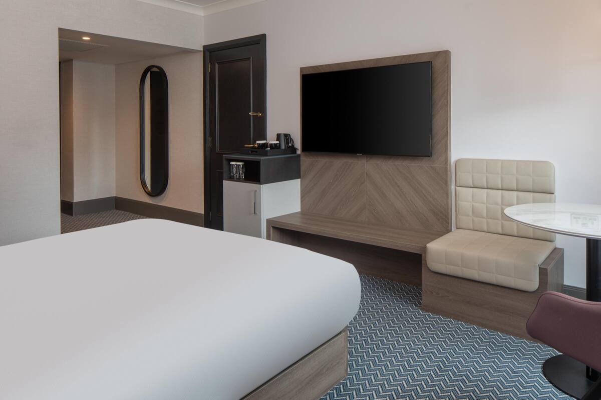 A hotel room with a large bed, a TV mounted on the wall, a small seating area with a cushioned bench, and a coffee machine on a cabinet.