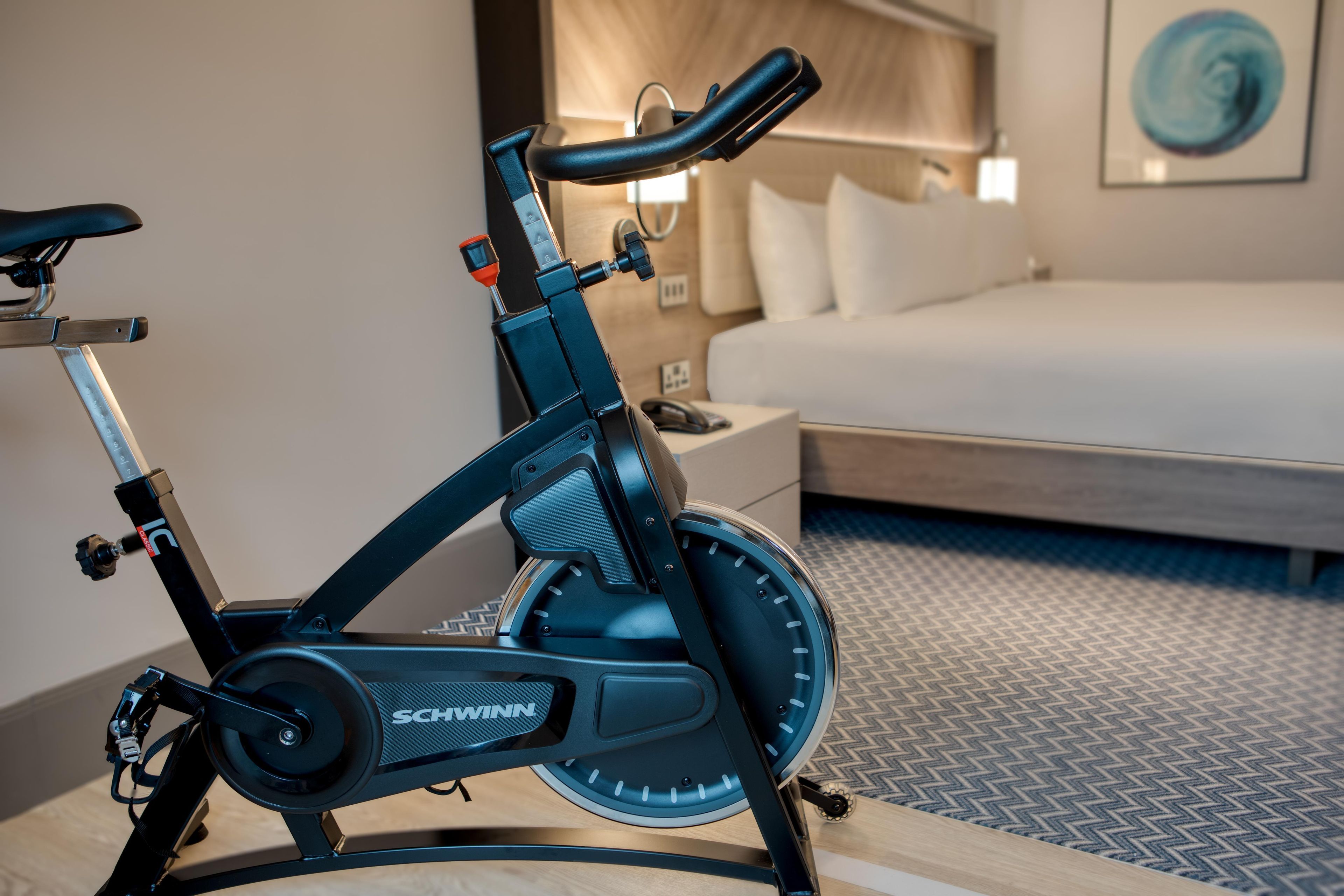 Una cyclette fissa in una camera d'albergo, con un letto e un comodino.