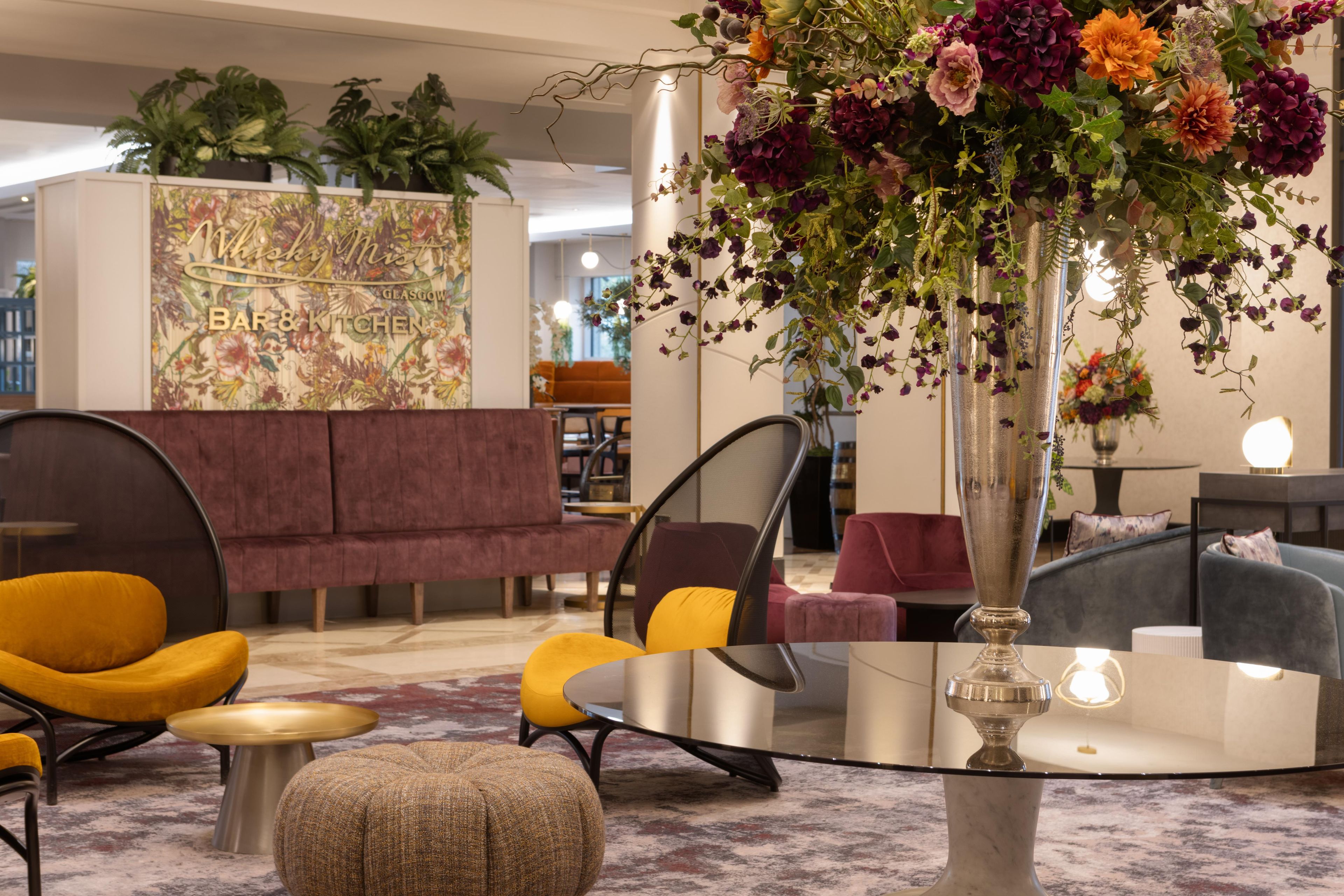Un área de recepción con arreglos de asientos, un jarrón decorativo con flores y una pared con un mural floral.