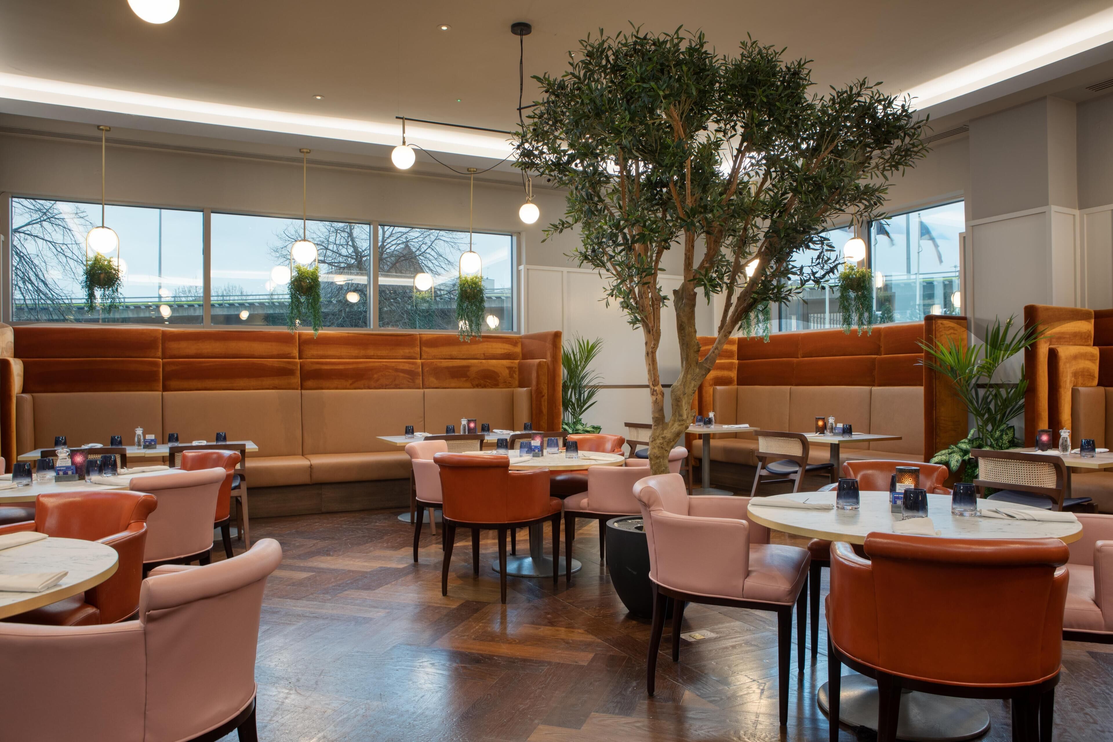 A restaurant with round tables, cushioned seating, pendant lighting, and large windows.