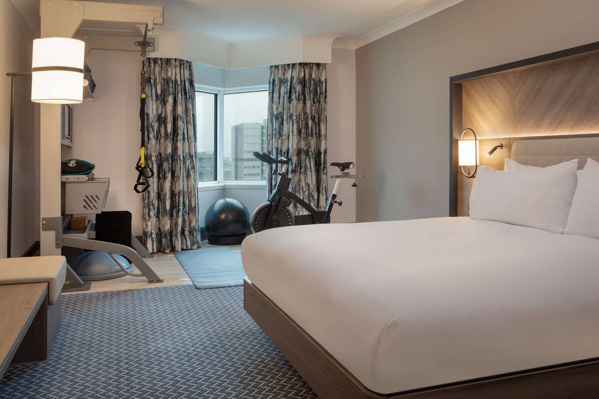 A hotel room with a large bed, exercise equipment, a desk, and a view of the city through the window.