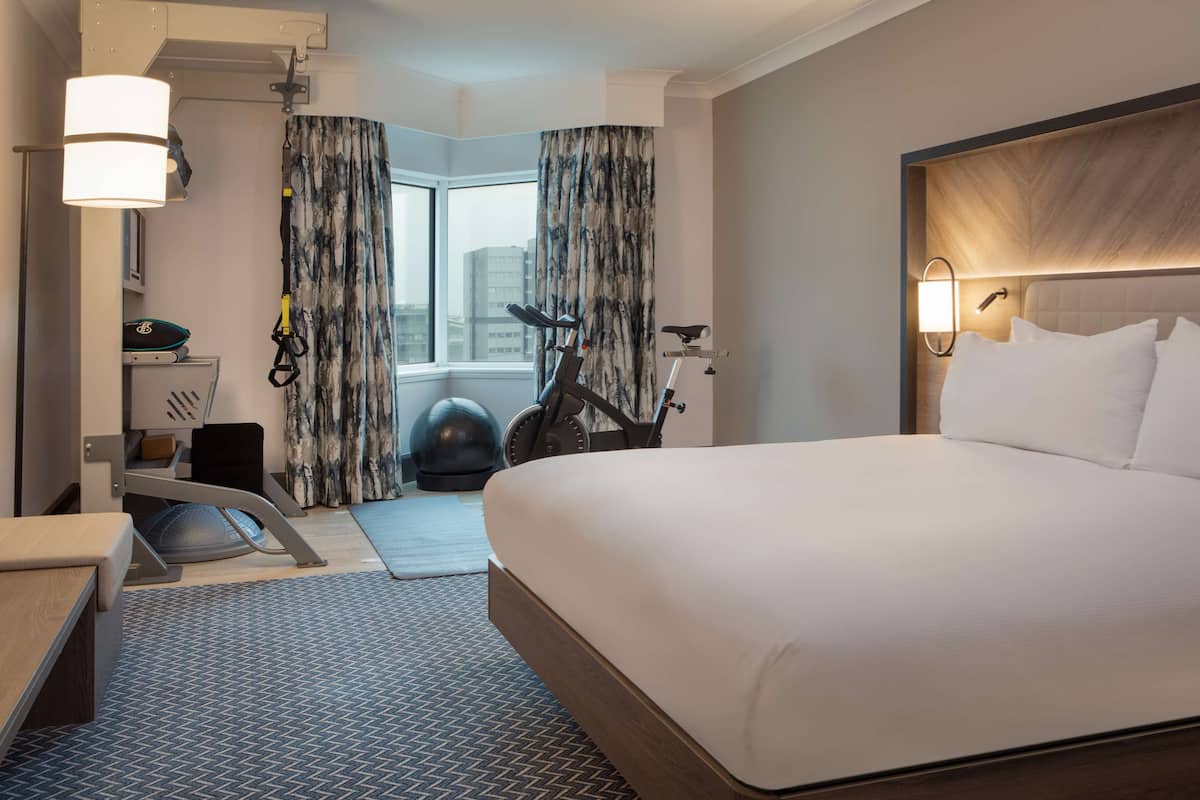 A hotel room with a large bed, exercise equipment, a desk, and a view of the city through the window.