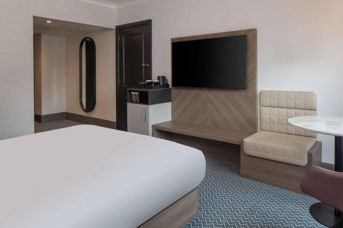 A hotel room with a large bed, a TV mounted on the wall, a small desk with a mirror, and a seating area with a cushioned bench and a round table.