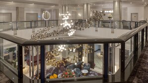 Un amplio y moderno vestíbulo de hotel con barandilla de cristal, una lámpara central y una zona de estar con sofás y una mesa de centro.