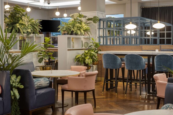 Relax in a stylish space with a round table, bar area, and comfortable seating, surrounded by lush greenery and a large window.