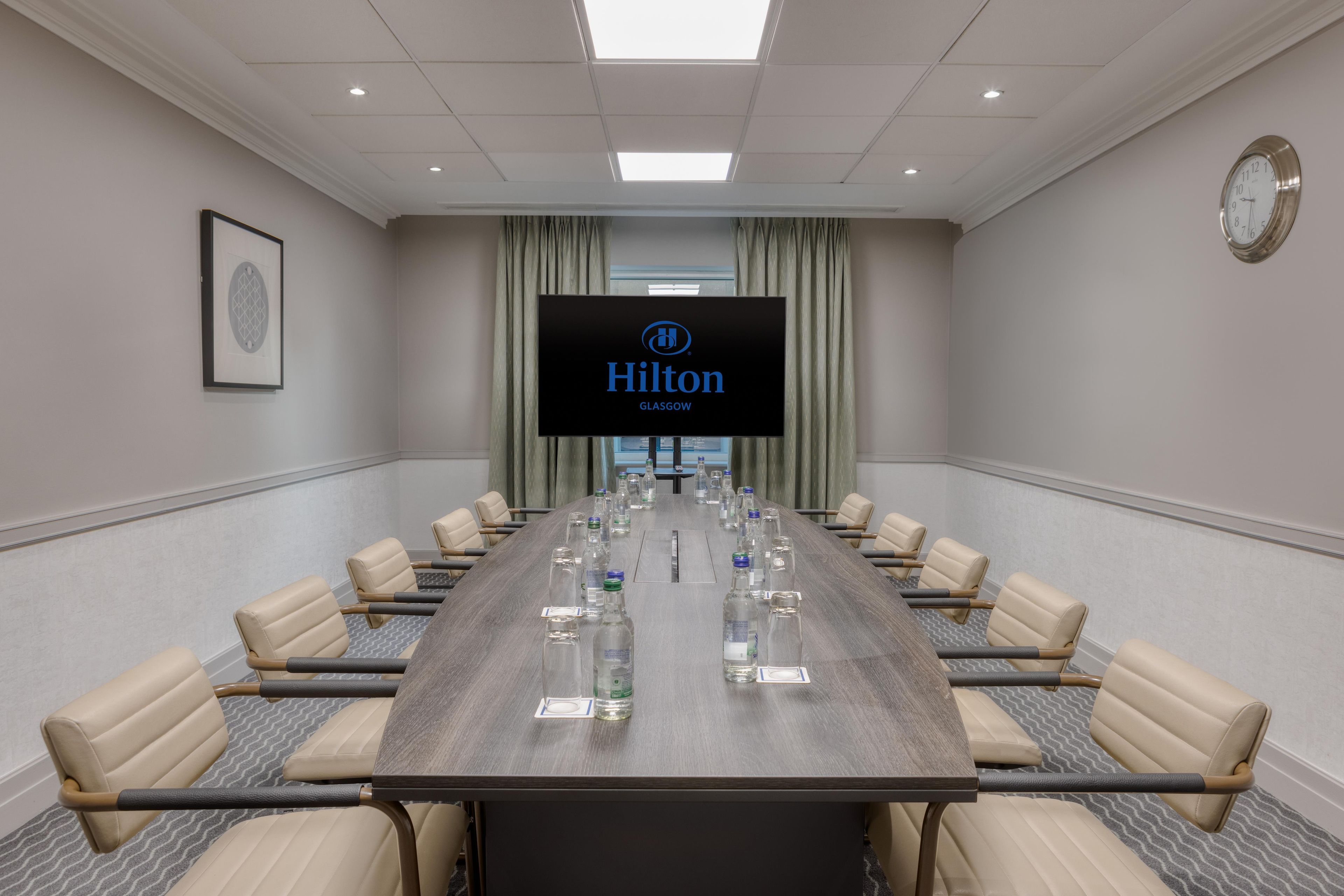 Een vergaderzaal met een lange tafel, stoelen, een scherm waarop 'Hilton Glasgow' staat en een klok aan de muur.