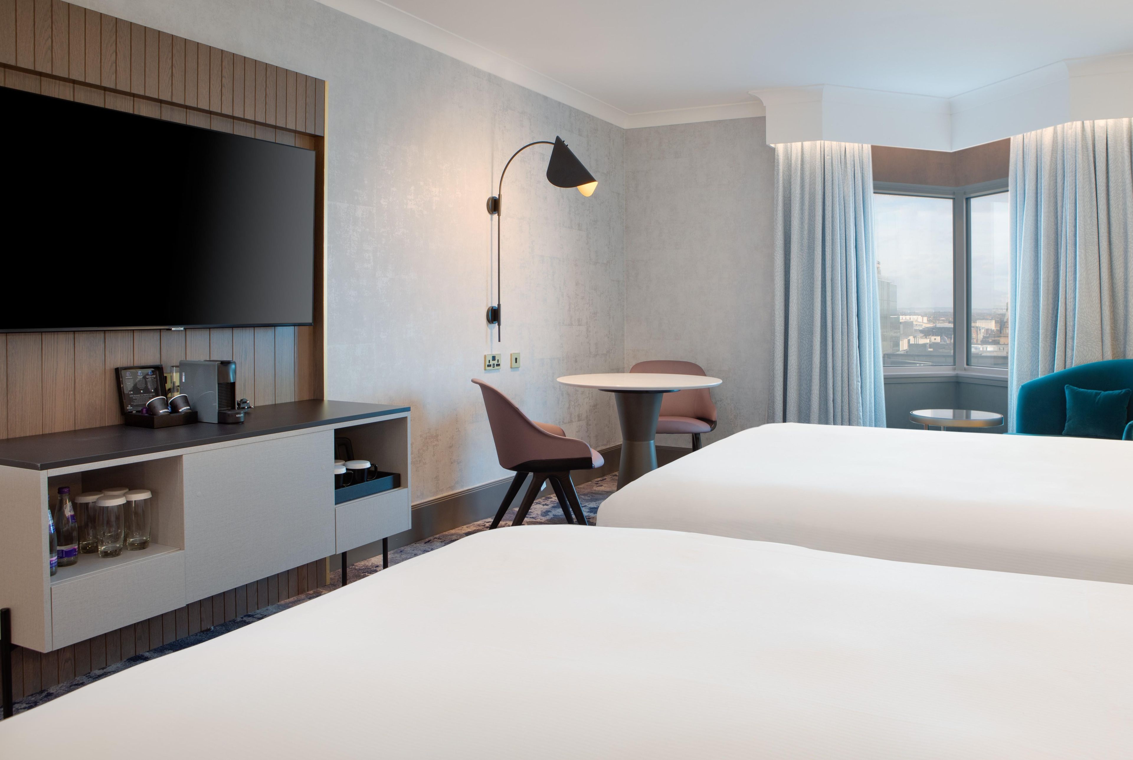 A hotel room with a flat-screen TV mounted on the wall, a desk with a lamp, a chair, a round table, and a bed.