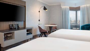A hotel room with a flat-screen TV mounted on the wall, a desk with a lamp, a chair, a round table, and a bed.