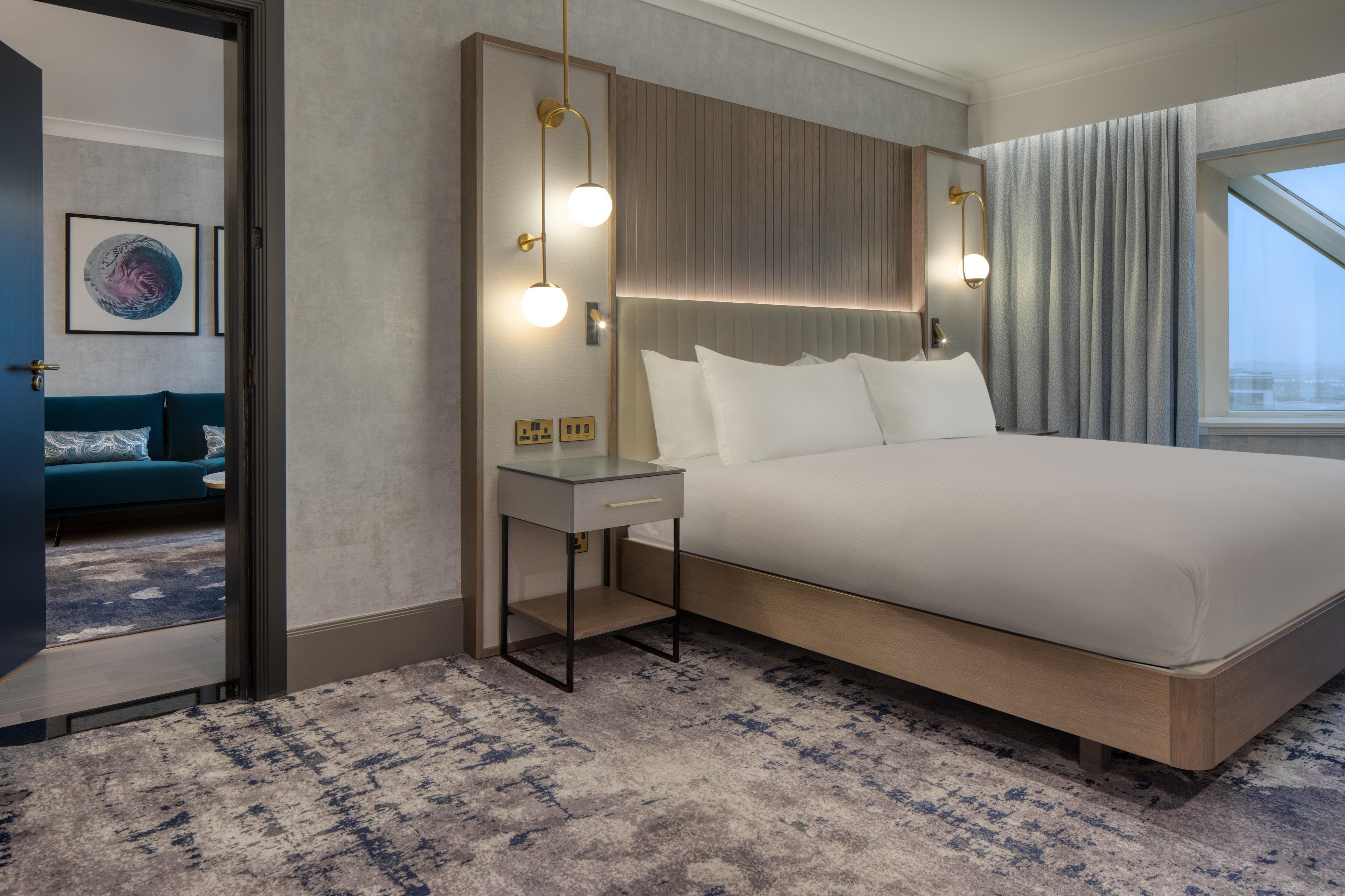 A modern hotel room with a large bed, bedside table, and a view of the city through the window.