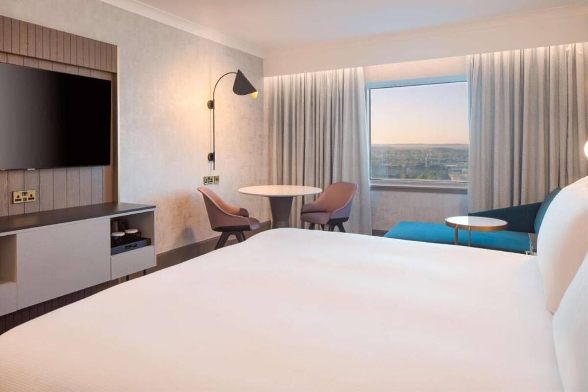 A hotel room with a bed, a TV mounted on the wall, a desk with a lamp, two chairs, a small table, and a view of the city through the window.