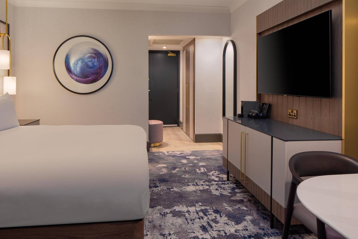 A hotel room with a bed, a desk with a flat-screen TV, a round painting, and a small stool.