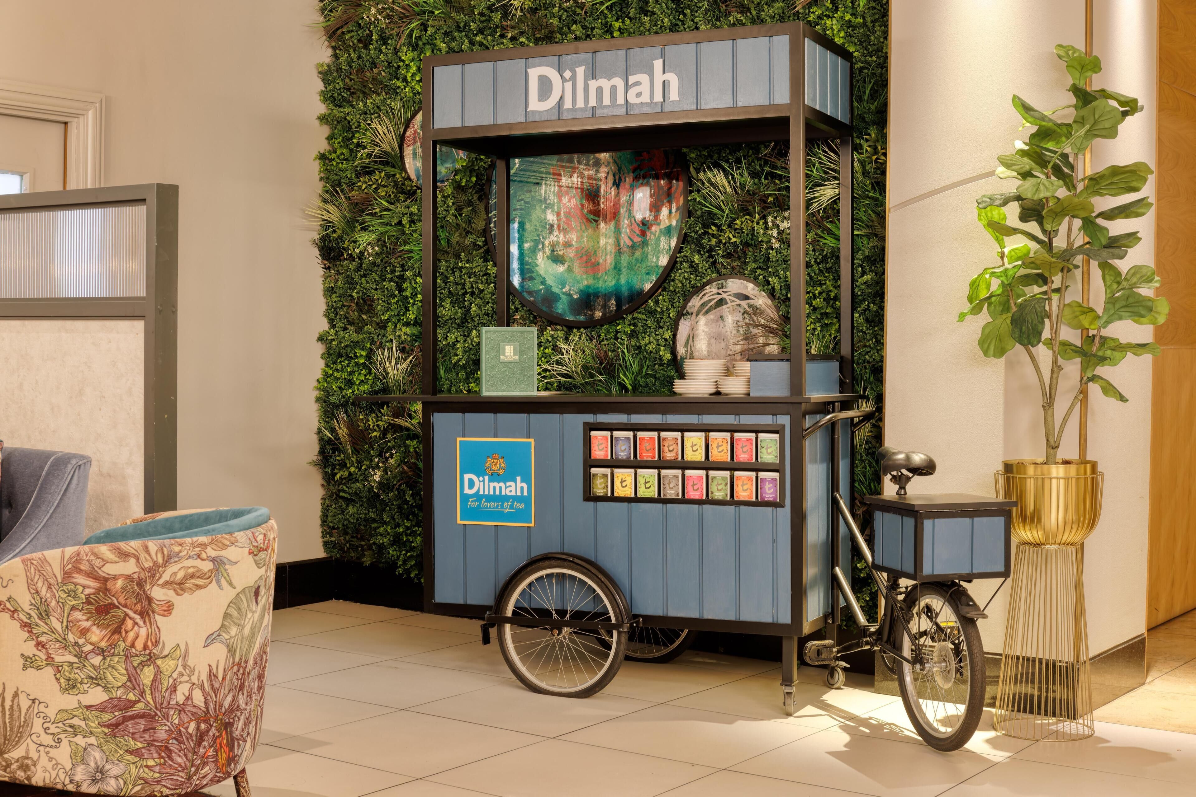 Un carrito de comida estacionario con el rótulo 'Dilmah', una variedad de productos envasados, una bicicleta y una planta en maceta.