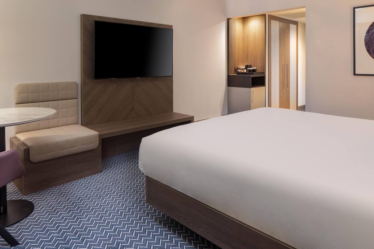 A hotel room with a bed, a TV mounted on the wall, a seating area with a table and chairs, and a small kitchenette.