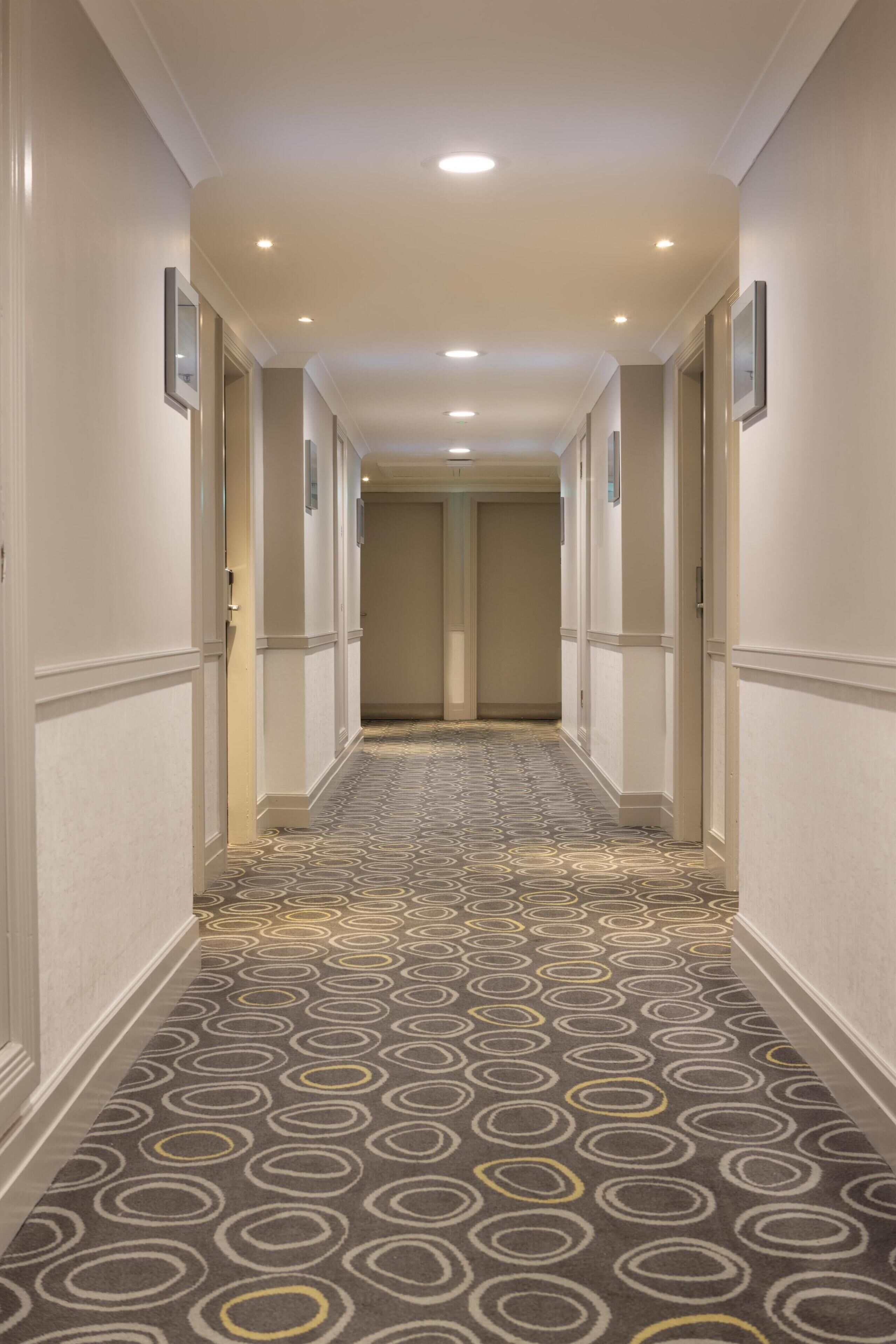 Ein Hotelgang mit gemustertem Teppich, weißen Wänden und Türen zu Zimmern.