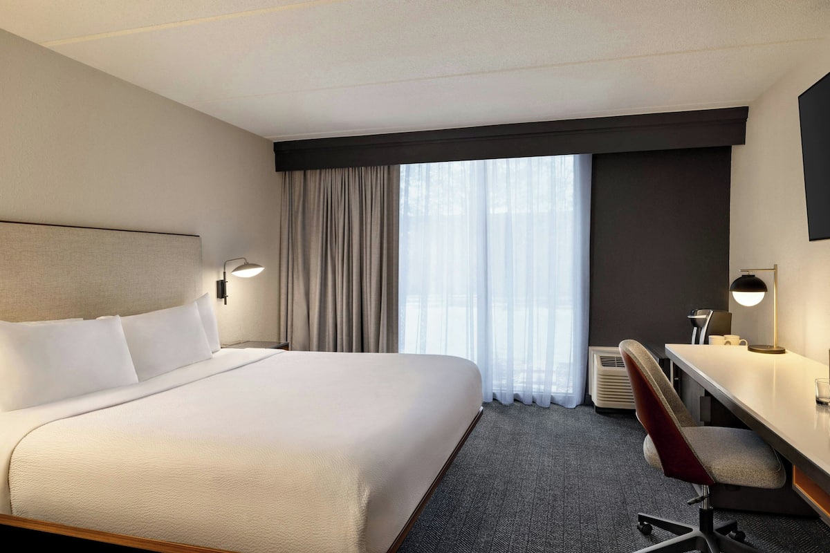 Habitación de hotel con una cama grande, un escritorio, una silla y un ventanal con cortinas.