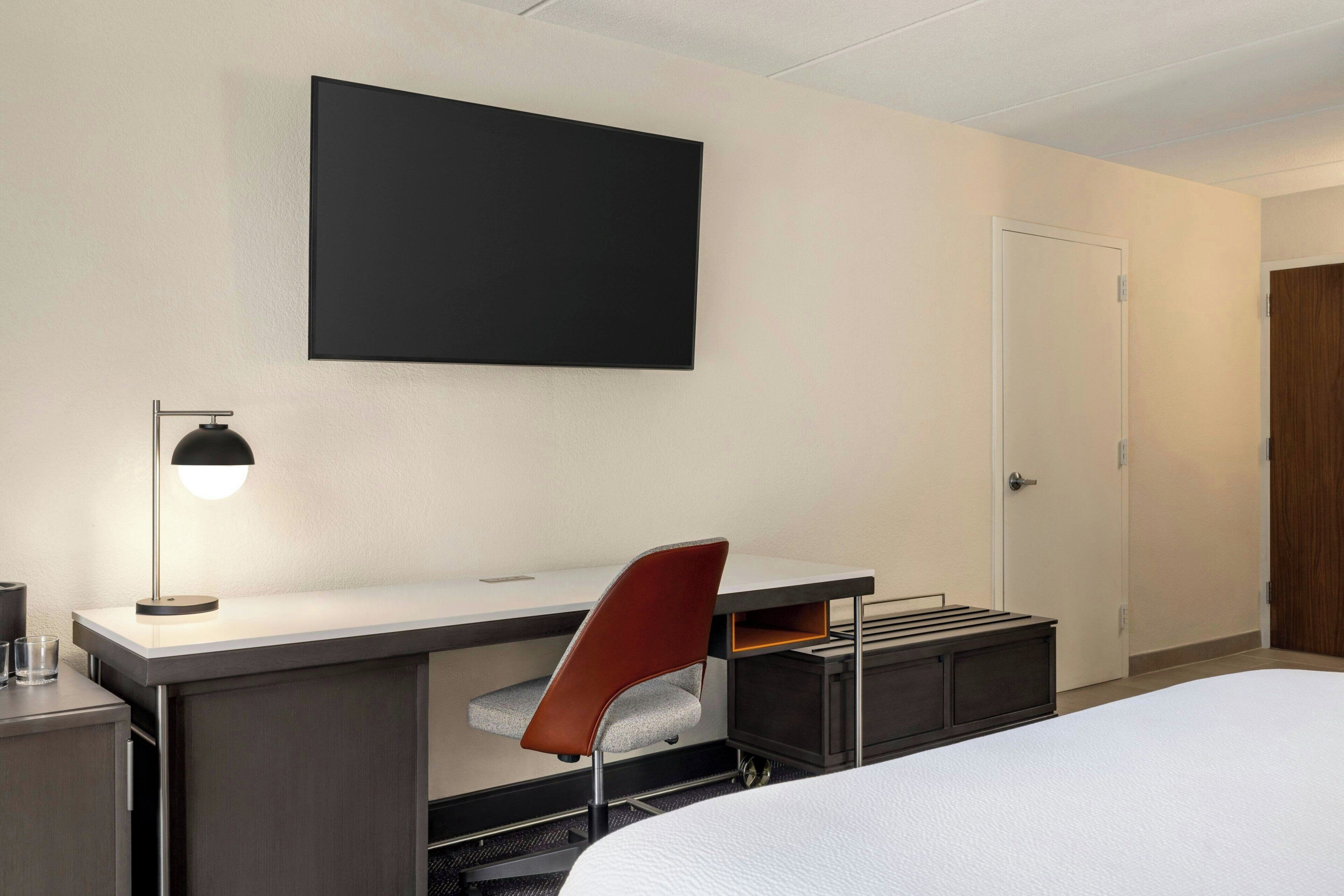 A hotel room with a bed, a desk with a chair, a TV mounted on the wall, and a door.