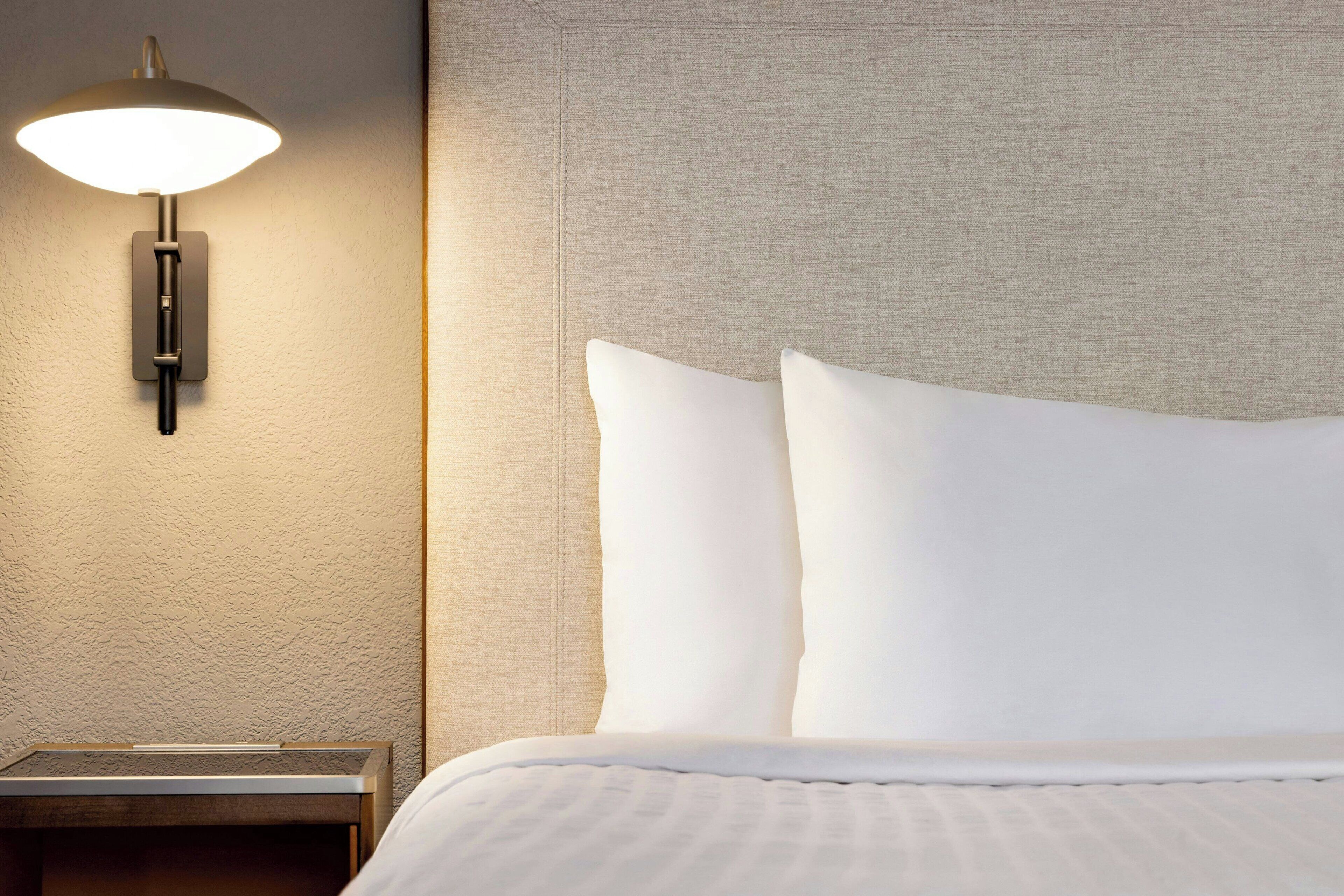 Una cama bien hecha con sábanas blancas y un cabecero, una lámpara de pared y una mesita de noche con una lámpara.