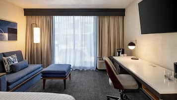 Habitación de hotel con sofá, escritorio, silla, mesita, lámpara y ventana grande con cortinas.
