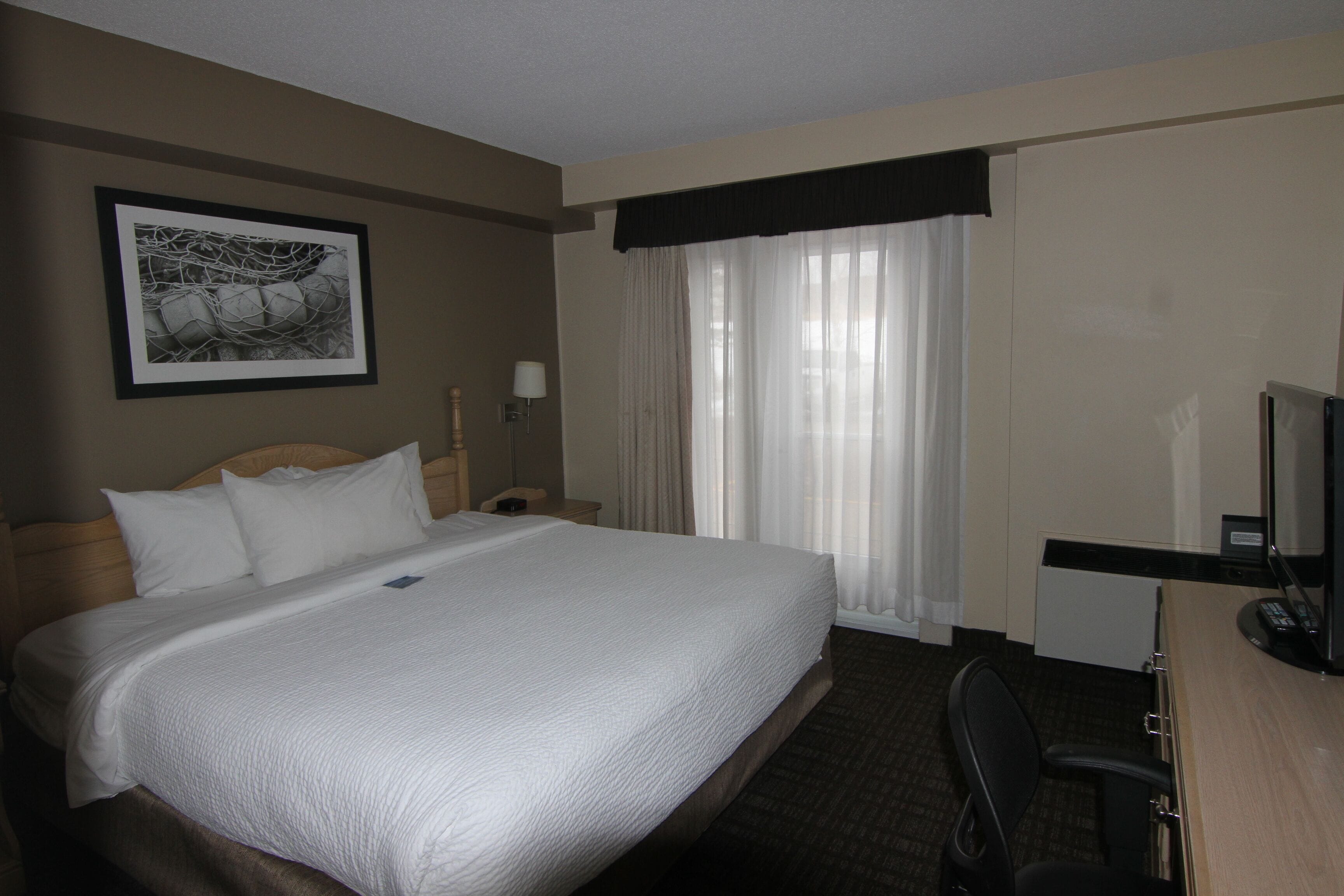 A hotel room with a large bed, a desk with a computer, a chair, a lamp, and a window with curtains.