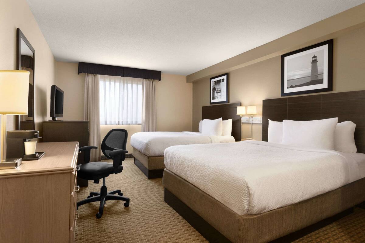 Quarto de hotel com duas camas, uma escrivaninha, uma cadeira, uma televisão e um quadro na parede.