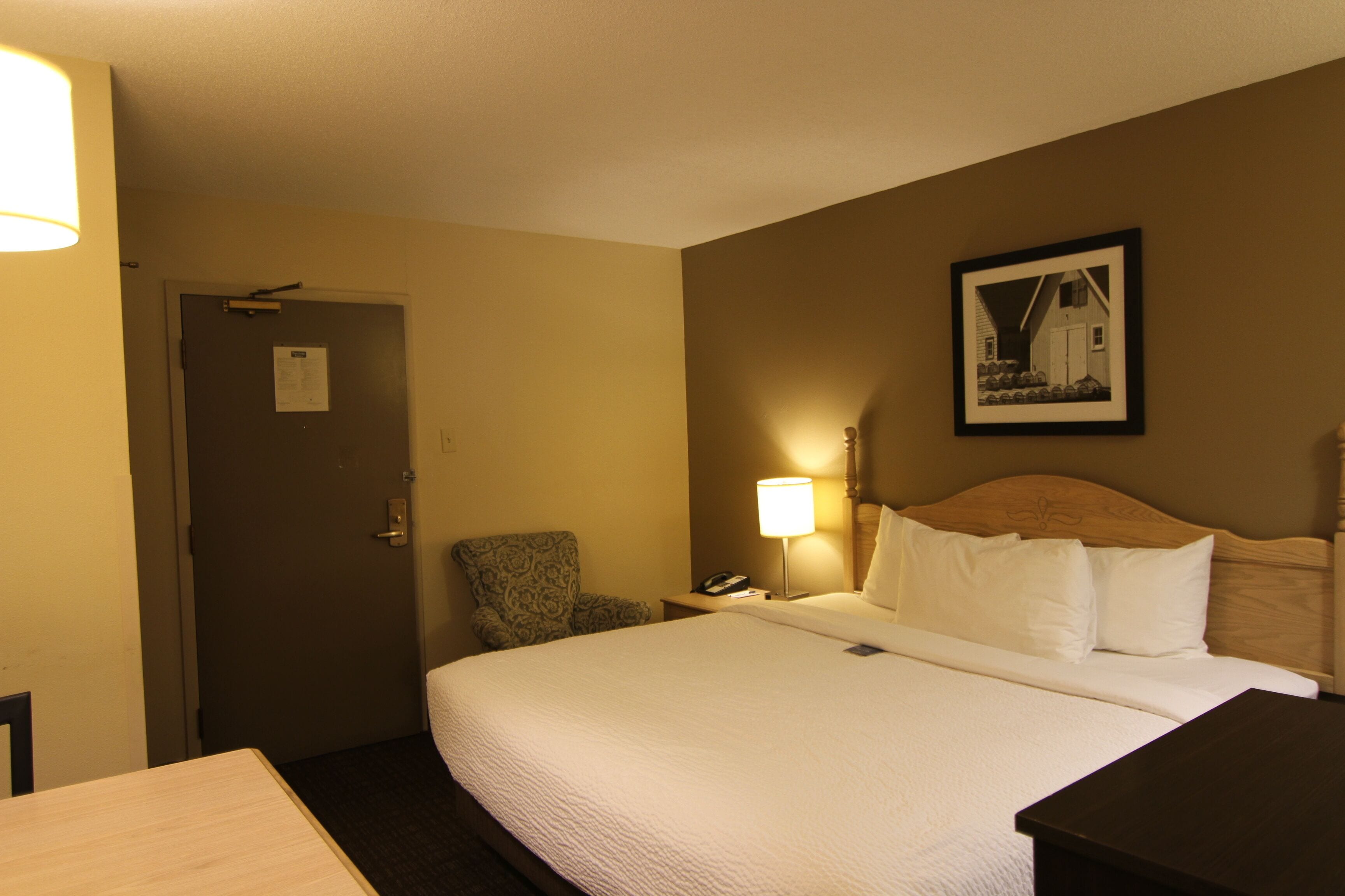 A hotel room with a large bed, a chair, a desk, a lamp, and a framed picture on the wall.