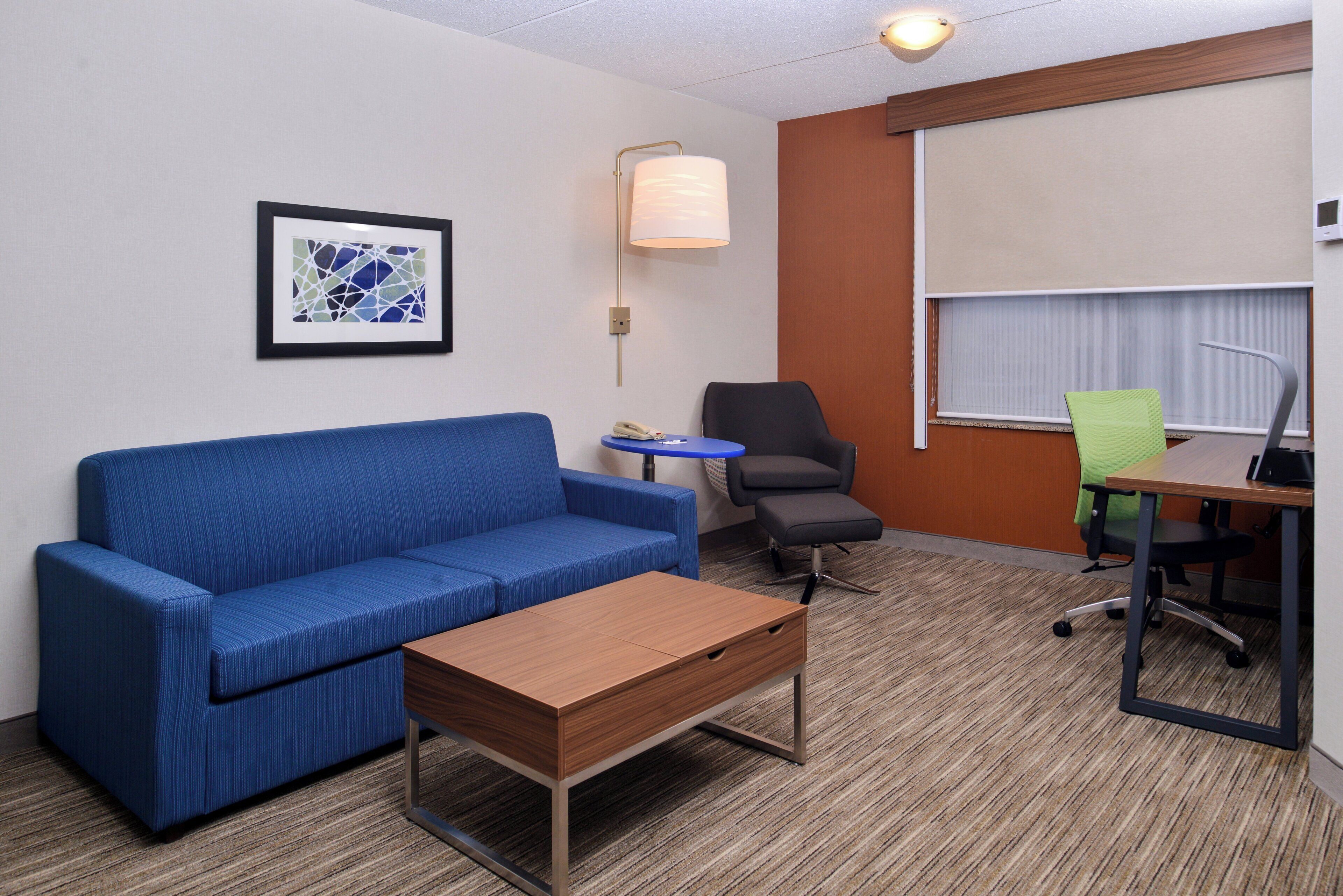 A hotel room with a blue sofa, a wooden coffee table, a small round table, a black office chair, and a green office chair.