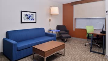 Une chambre d’hôtel avec un canapé bleu, une table basse en bois, une petite table ronde, un fauteuil de bureau noir et un fauteuil de bureau vert.