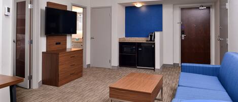 Una habitación de hotel moderna con un aparador de madera, un televisor de pantalla plana, una kitchenette, un sofá azul y una pequeña mesa.