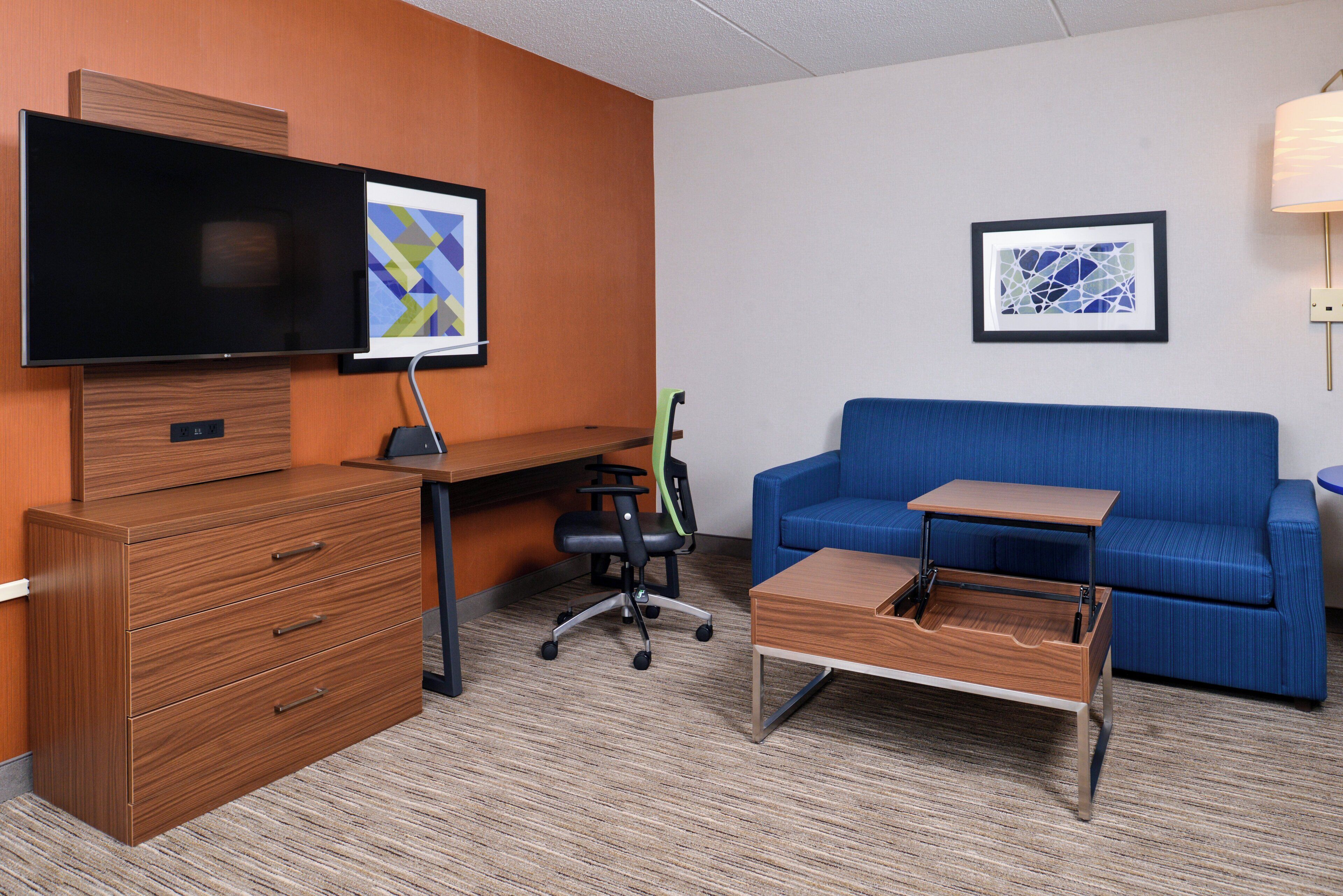 A hotel room with a wooden desk, a television, a blue sofa, and a small wooden table.