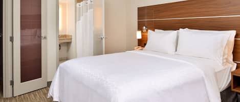 A hotel room with a large bed, a wooden headboard, a bedside table with a lamp, and a bathroom visible through an open door.