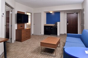 Habitación de hotel con televisor de pantalla plana, cómoda de madera, sofá azul, una mesita pequeña y una kitchenette con cafetera.