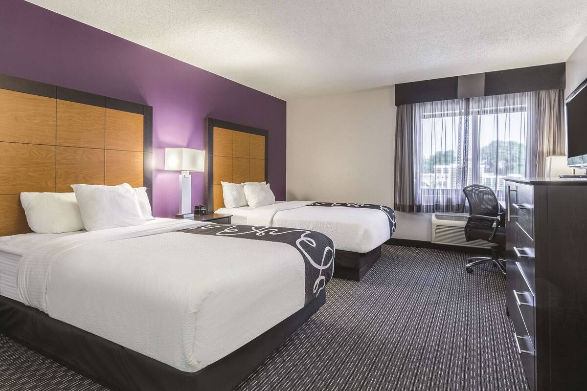 A hotel room with two beds, a desk with a chair, a TV, and a window with curtains.