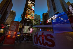 Uma rua urbana movimentada com edifícios altos, outdoors e um ônibus com a inscrição 'tkts'.