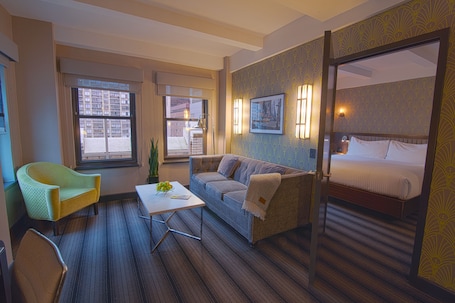 A hotel room with a bed, a sofa, a chair, a small table, and two windows.. Hotel Edison Times Square