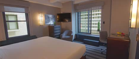 Habitación de hotel con una cama grande, un escritorio, una silla, un televisor y ventanas con vista a la ciudad.
