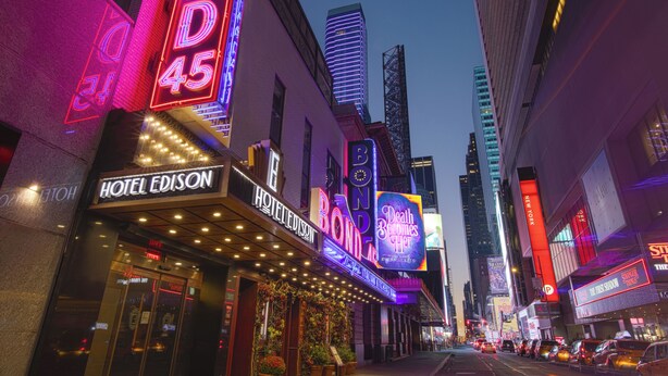 A hotel with a neon sign, a theater marquee, and a tall building in the background.