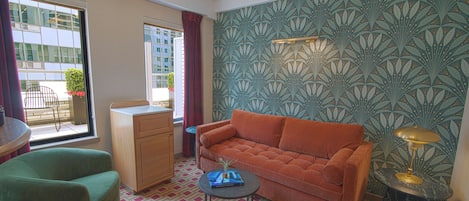 Una habitación con sofá, sillón, mesita, lámpara, ventana con cortinas y un papel tapiz estampado.