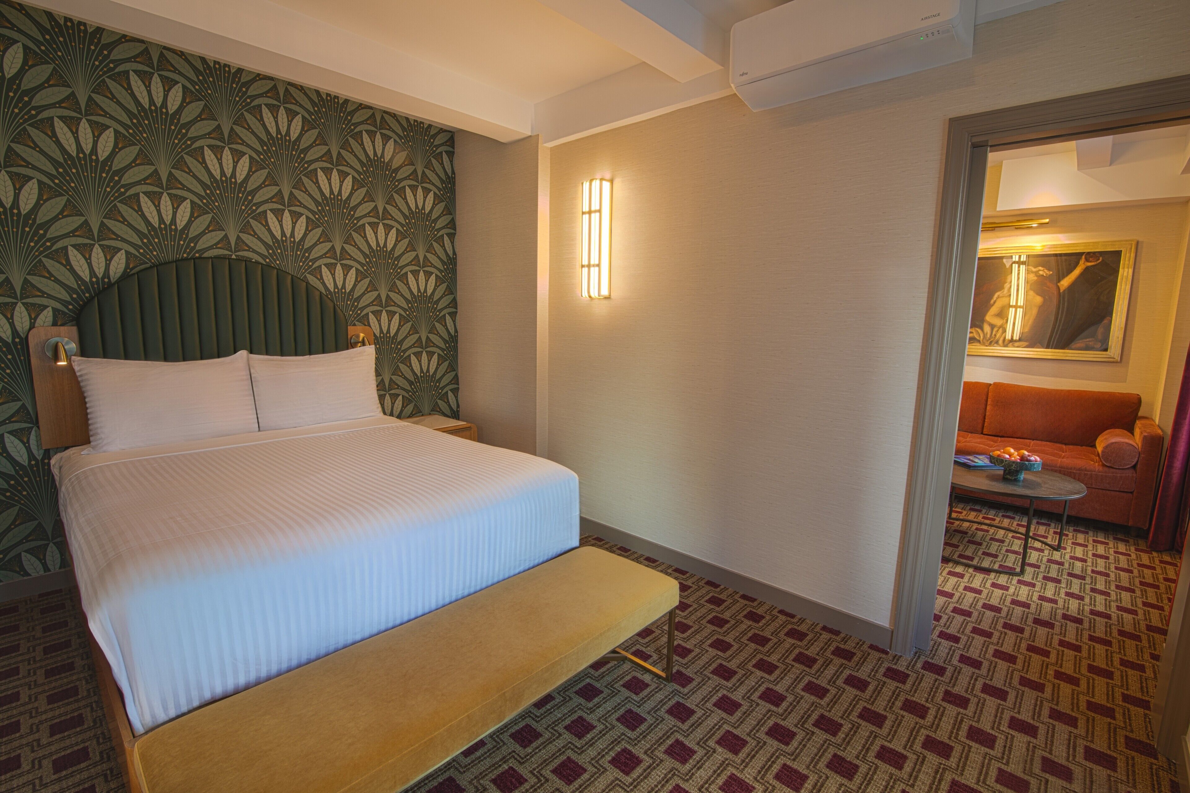 A hotel room with a bed, bench, patterned carpet, wall art, and a sofa.