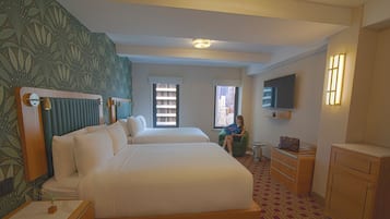 Ein Hotelzimmer mit zwei Betten, einem Fernseher, einem Schreibtisch und einem Fenster mit Blick auf die Stadt.