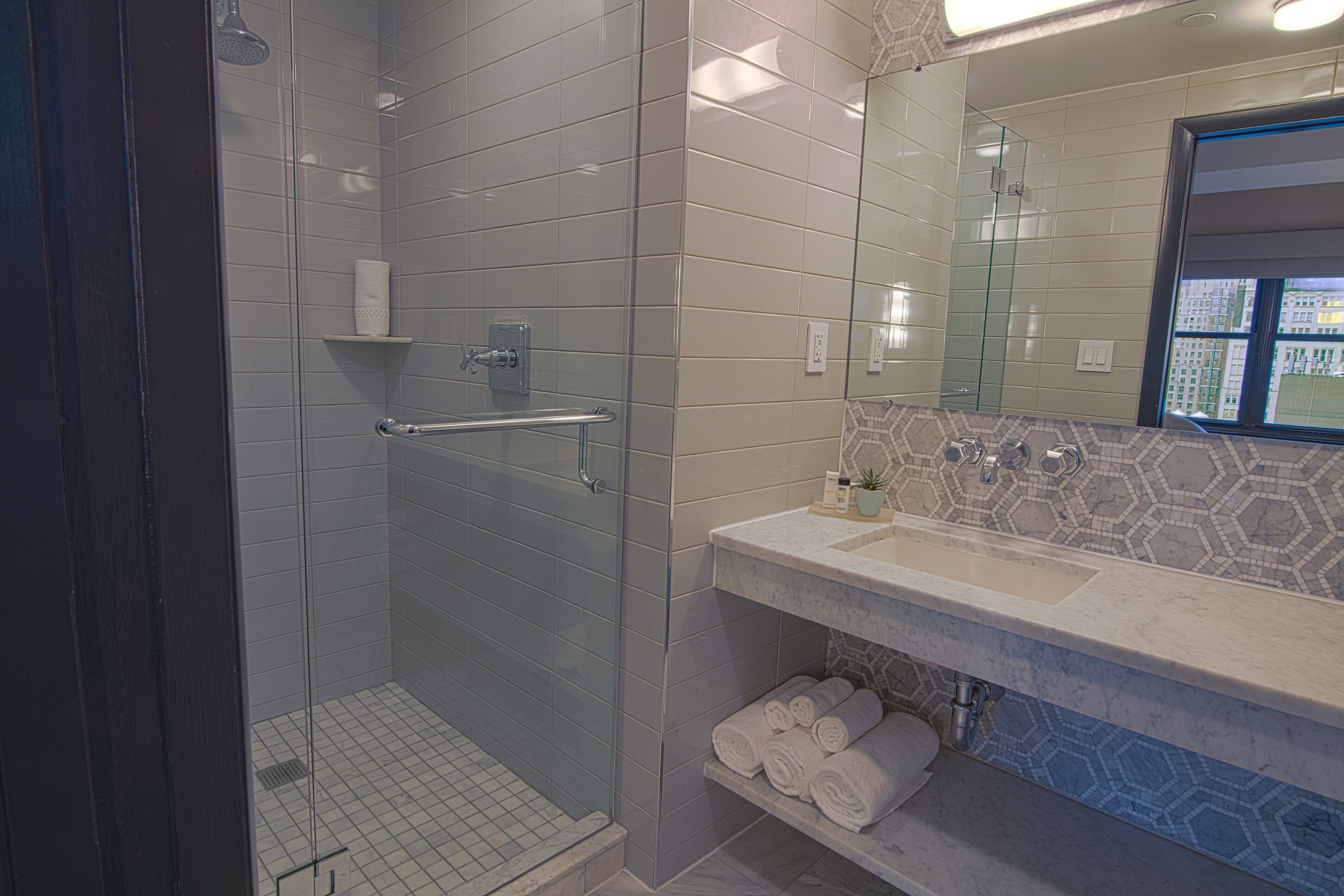 A modern bathroom with a glass-enclosed shower, a vanity with a marble countertop, and a view of buildings through a window.