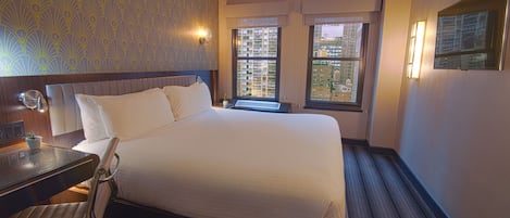 Habitación de hotel con una cama grande, un escritorio con una silla, una mesita con una planta y dos ventanas con vistas a la ciudad.