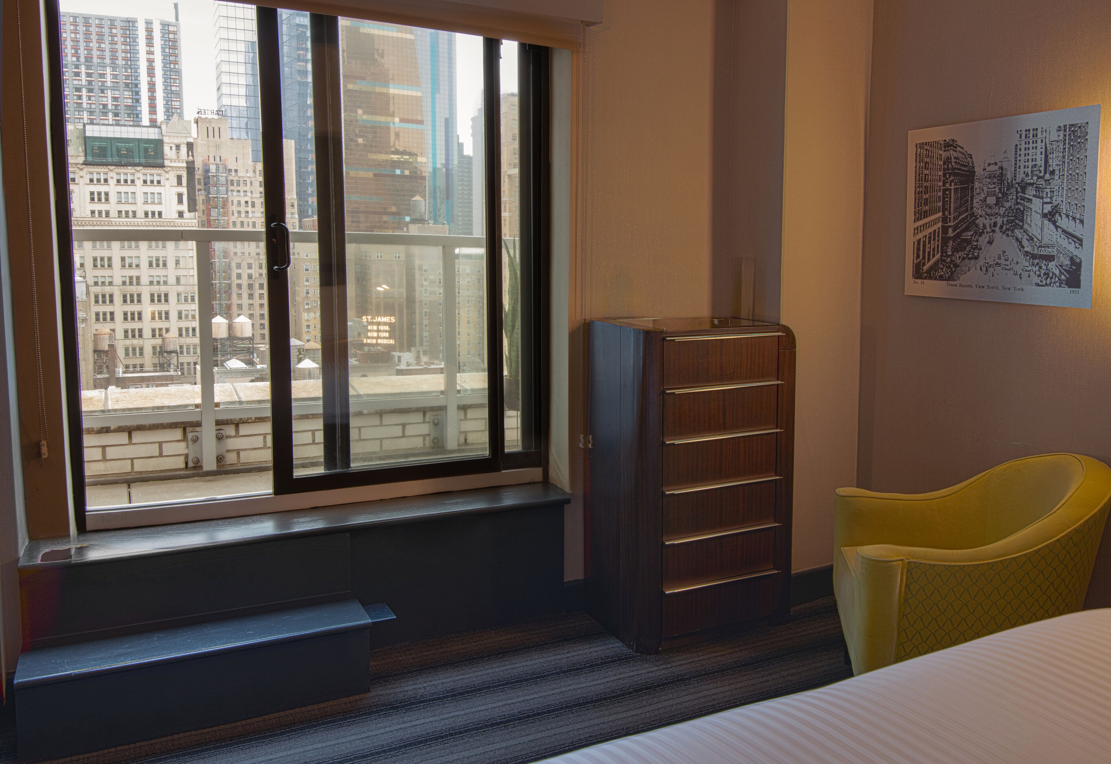 A hotel room with a large window offering a city view, a wooden bookshelf, a yellow chair, and a bed.