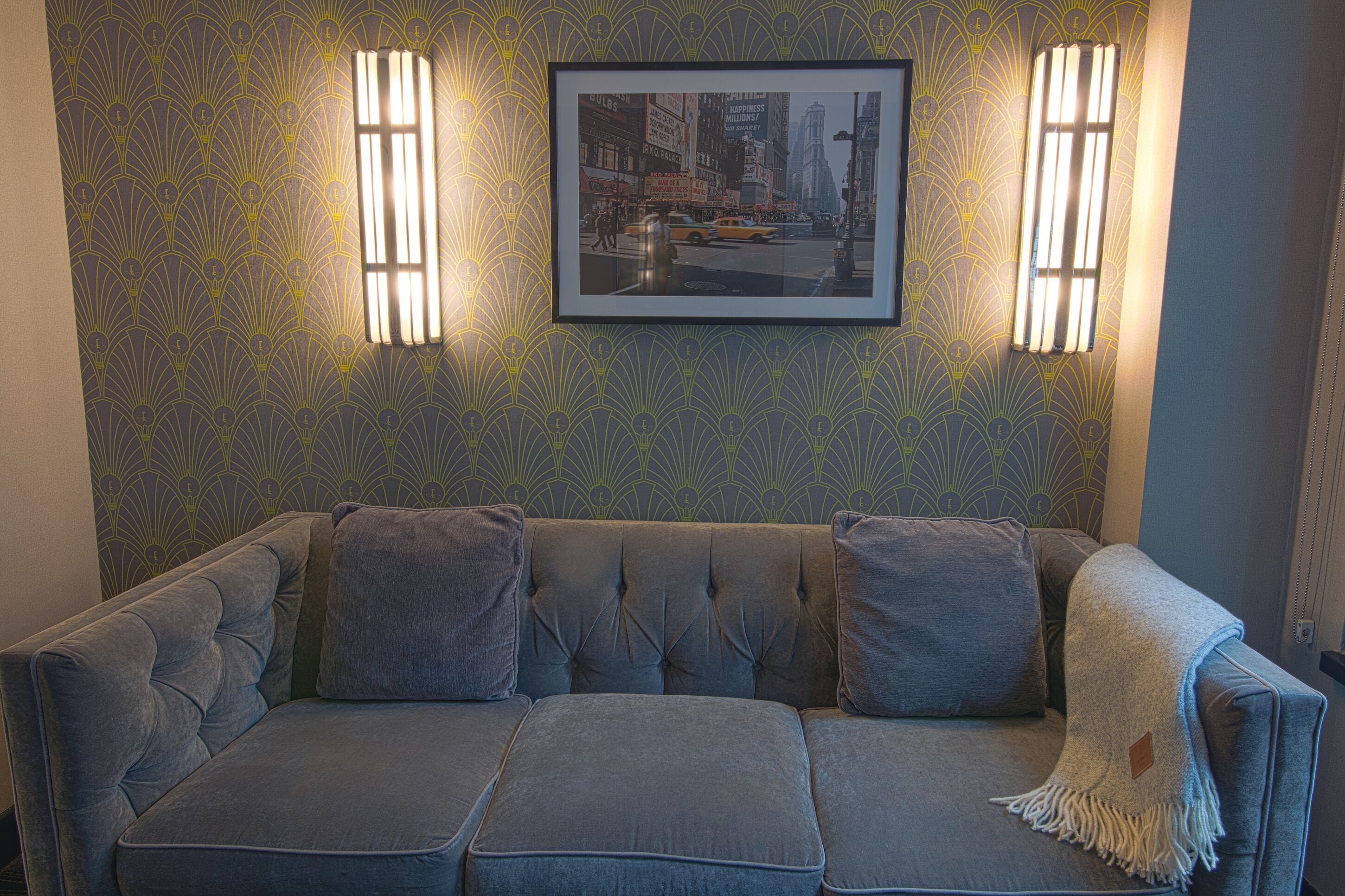 A gray sofa with a tufted backrest, a framed picture on the wall, and two wall-mounted light fixtures.