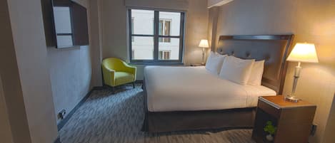 A hotel room with a large bed, two bedside lamps, a small table with a plant, a chair, and a window with a view of buildings.