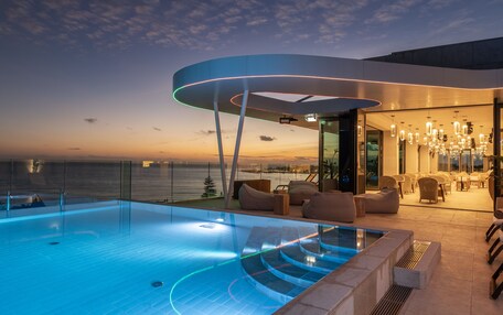 A modern rooftop pool area with a view of the ocean at sunset, featuring a glass-enclosed lounge and outdoor seating.