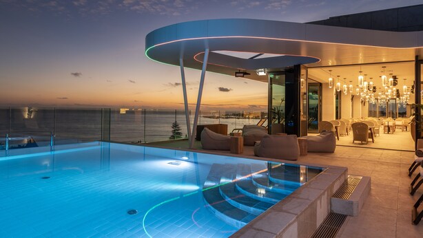 Un área de piscina moderna en la azotea con vistas al océano al atardecer, que cuenta con un salón con paredes de vidrio y asientos al aire libre.