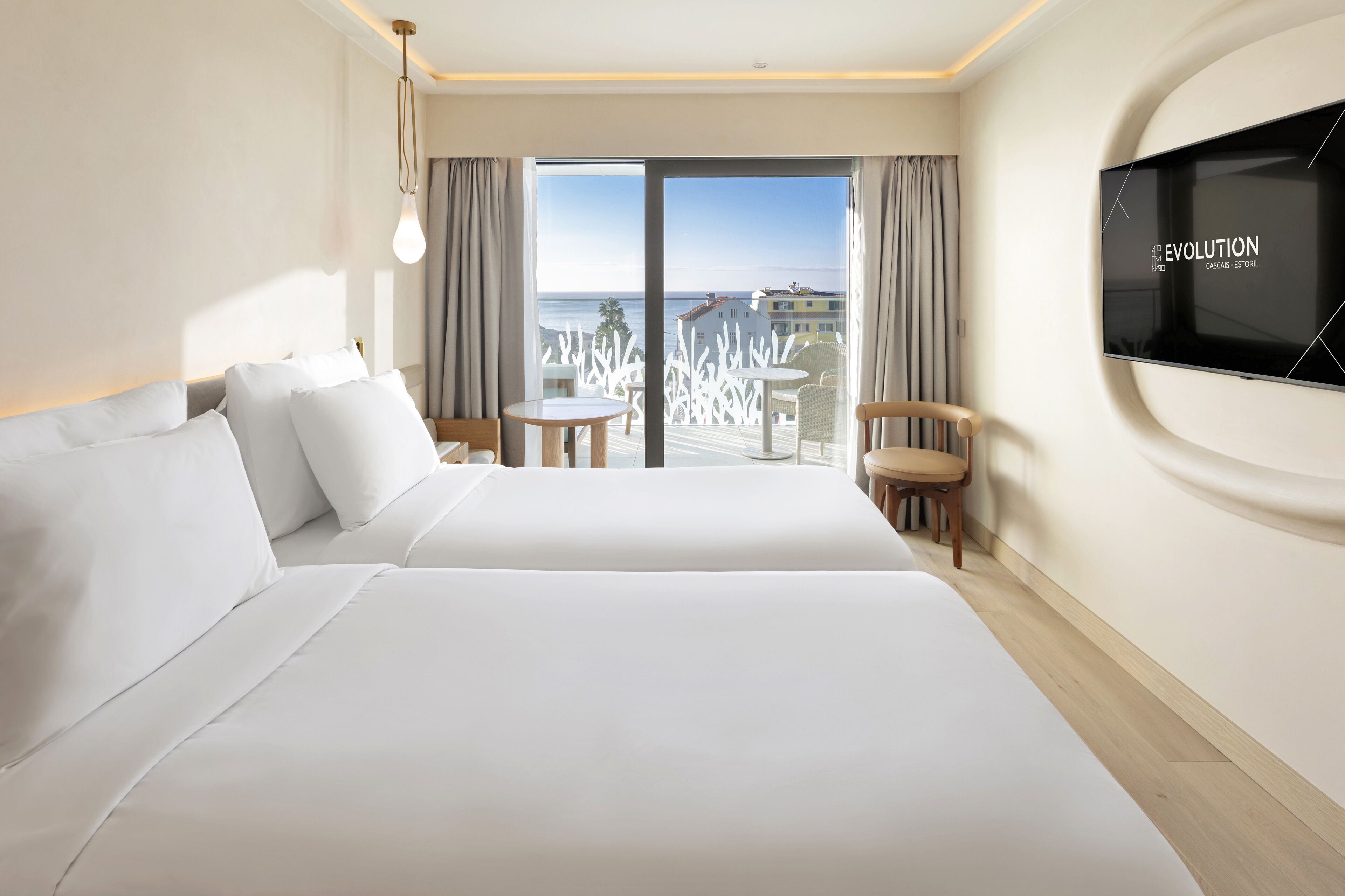 Habitación de hotel con una cama grande, un televisor montado en la pared, un balcón con vistas al mar y una pequeña mesa con sillas.