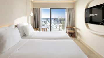 Habitación de hotel con una cama grande, un televisor montado en la pared, un balcón con vistas al mar y una pequeña mesa con sillas.