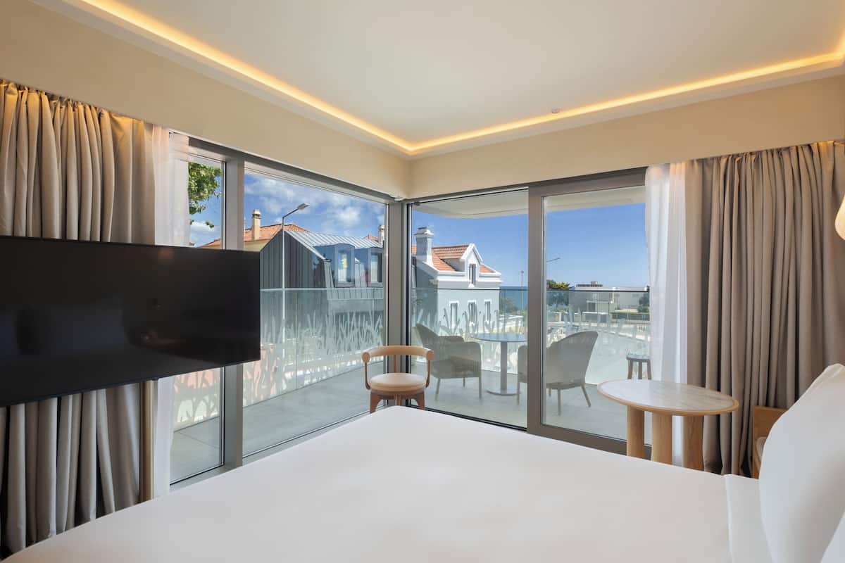A modern hotel room with a large bed, a flat-screen TV, a balcony with a glass door, and a view of buildings and a clear sky.